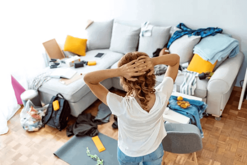 Woman in cluttered room facing away, hands on head