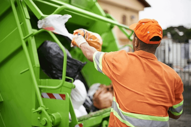 Worker in orange loading trash into green garbage truck